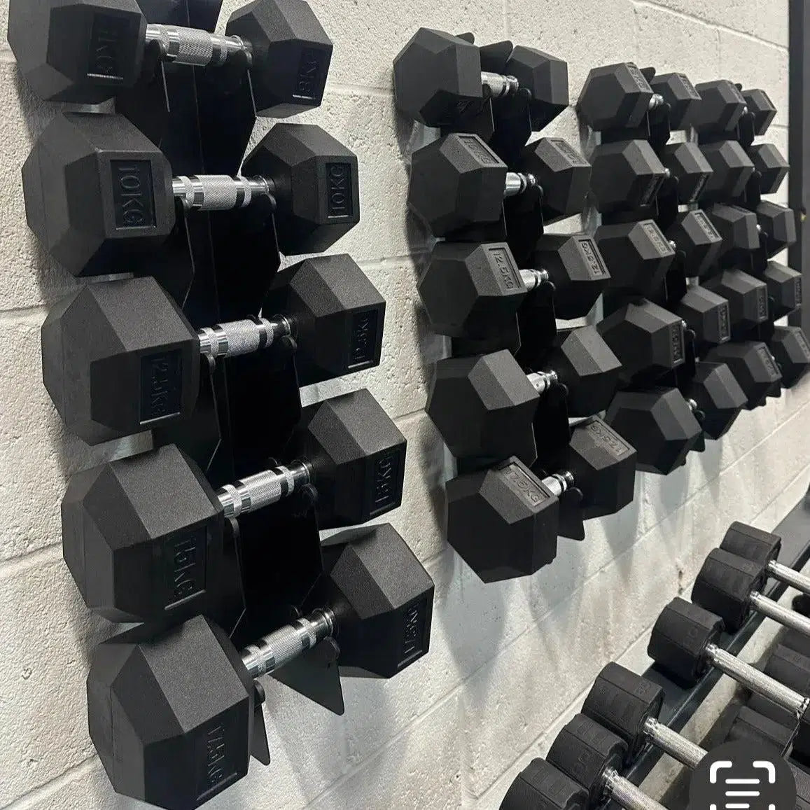 Hex dumbbell sets in varying weights neatly organized on two wall-mounted black metal racks against a gray wall.