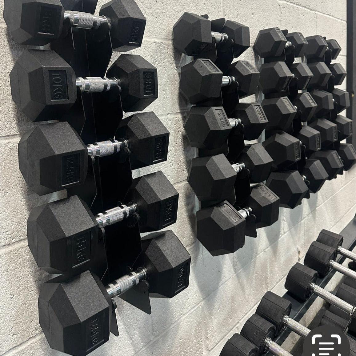 Black hex dumbbells neatly arranged on two wall-mounted metal racks against a white brick wall.